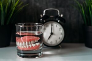 Set of dentures in a glass with clear liquid next to an alarm clock